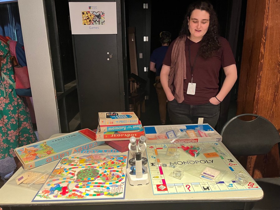 A Museum staff member stands behind a table showcasing vintage board games including Candy Land and Monopoly.