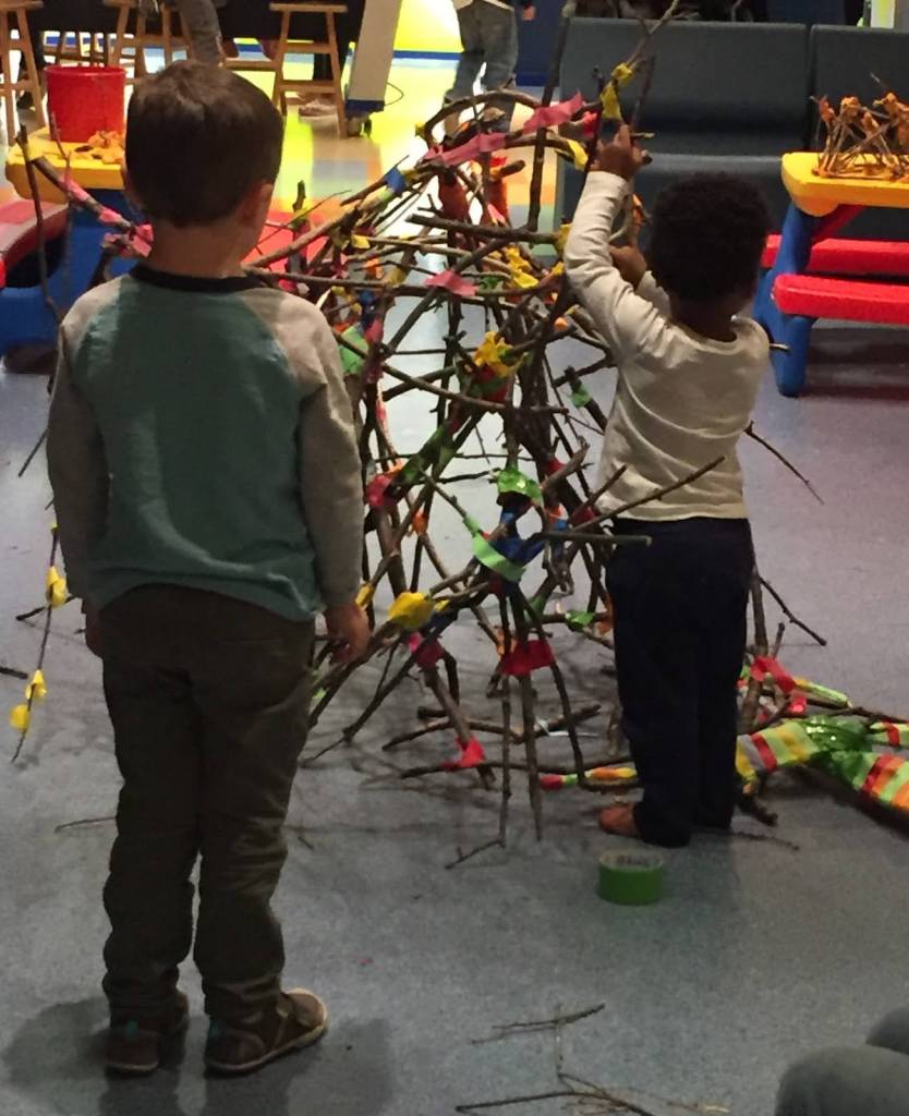 A young child wraps a piece of tape around a twig to secure it in place. Another child stands a few steps behind, watching how the tape is applied to the larger twig structure.